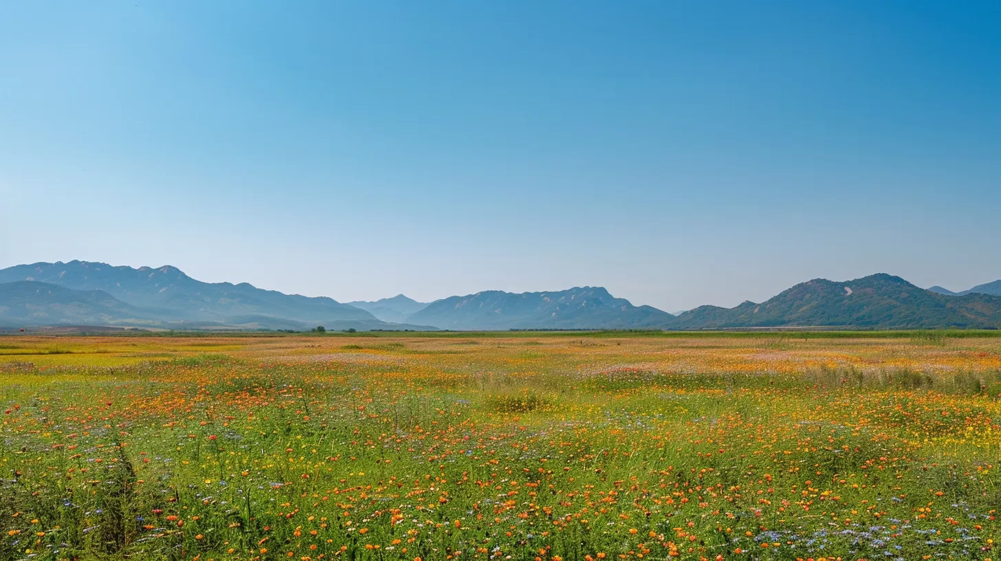 a serene, sunlit landscape featuring a vibrant, blossoming field of wildflowers stretching towards a distant, majestic mountain range under a clear blue sky.